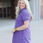 Woman with blonde hair standing in a side profile position wearing the Annie Dress in a lifestyle scene. The focal point of this image is the purple denim, pleating on sleeves as well as cuff. And folded collar. 