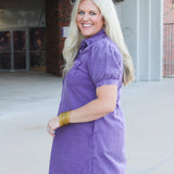 Woman with blonde hair standing in a side profile position wearing the Annie Dress in a lifestyle scene. The focal point of this image is the purple denim, pleating on sleeves as well as cuff. And folded collar. 