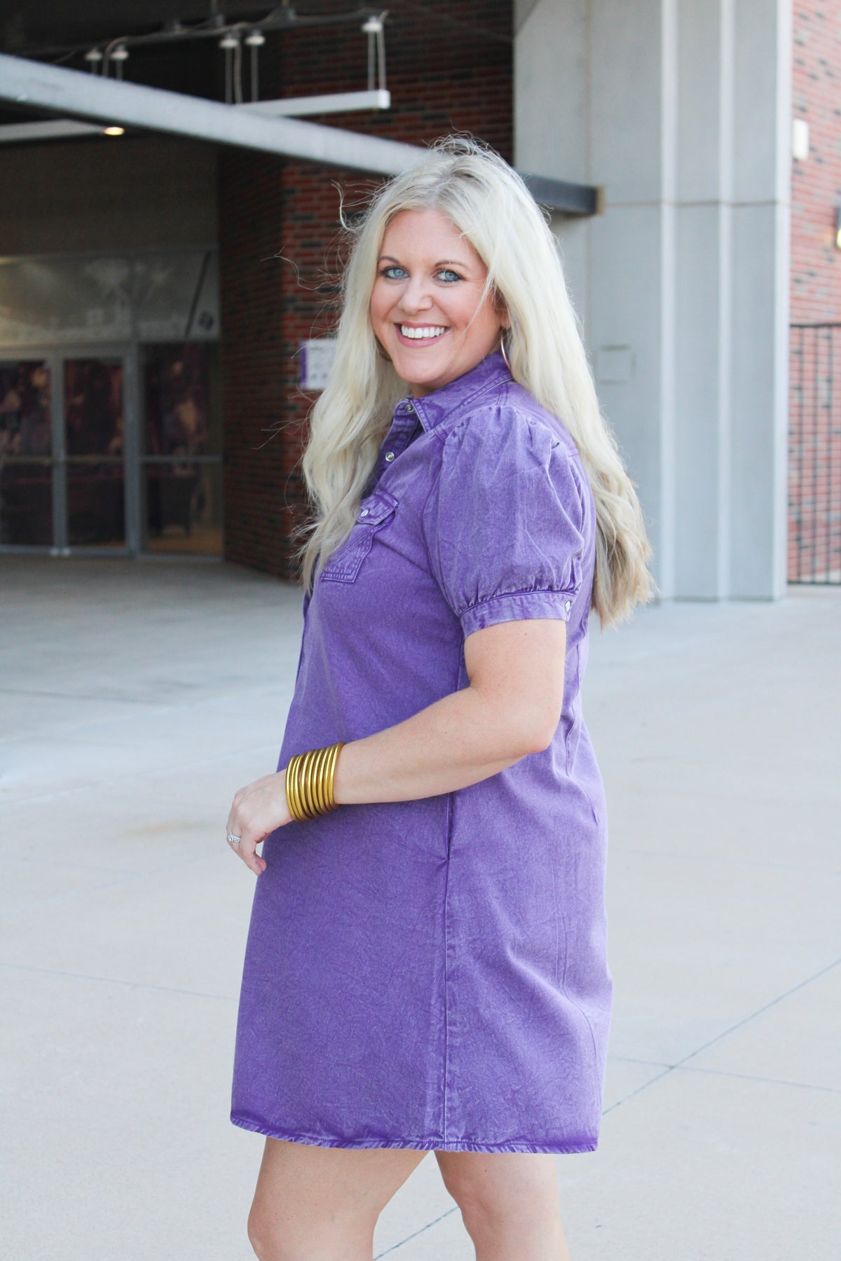 Woman with blonde hair standing in a side profile position wearing the Annie Dress in a lifestyle scene. The focal point of this image is the purple denim, pleating on sleeves as well as cuff. And folded collar. 
