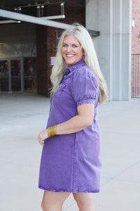 Woman with blonde hair standing in a side profile position wearing the Annie Dress in a lifestyle scene. The focal point of this image is the purple denim, pleating on sleeves as well as cuff. And folded collar. 