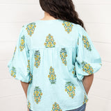 Woman with dark hair standing in a back profile position wearing the Ava Top against a white background. The focal point of this image is the yellow floral pattern as well as pleating on back and ruffle sleeve. The model paired this top with dark-wash denim jeans. 