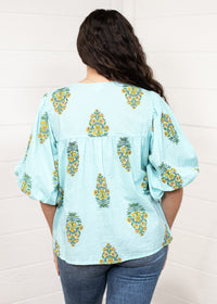 Woman with dark hair standing in a back profile position wearing the Ava Top against a white background. The focal point of this image is the yellow floral pattern as well as pleating on back and ruffle sleeve. The model paired this top with dark-wash denim jeans. 