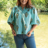 Woman with red hair standing in a front profile position wearing the Ava Top in a lifestyle scene. The focal point of this image is distressed fabric with same graphics along the chest, v-neckline, and tassels. The model paired this top with dark-wash denim jeans. 