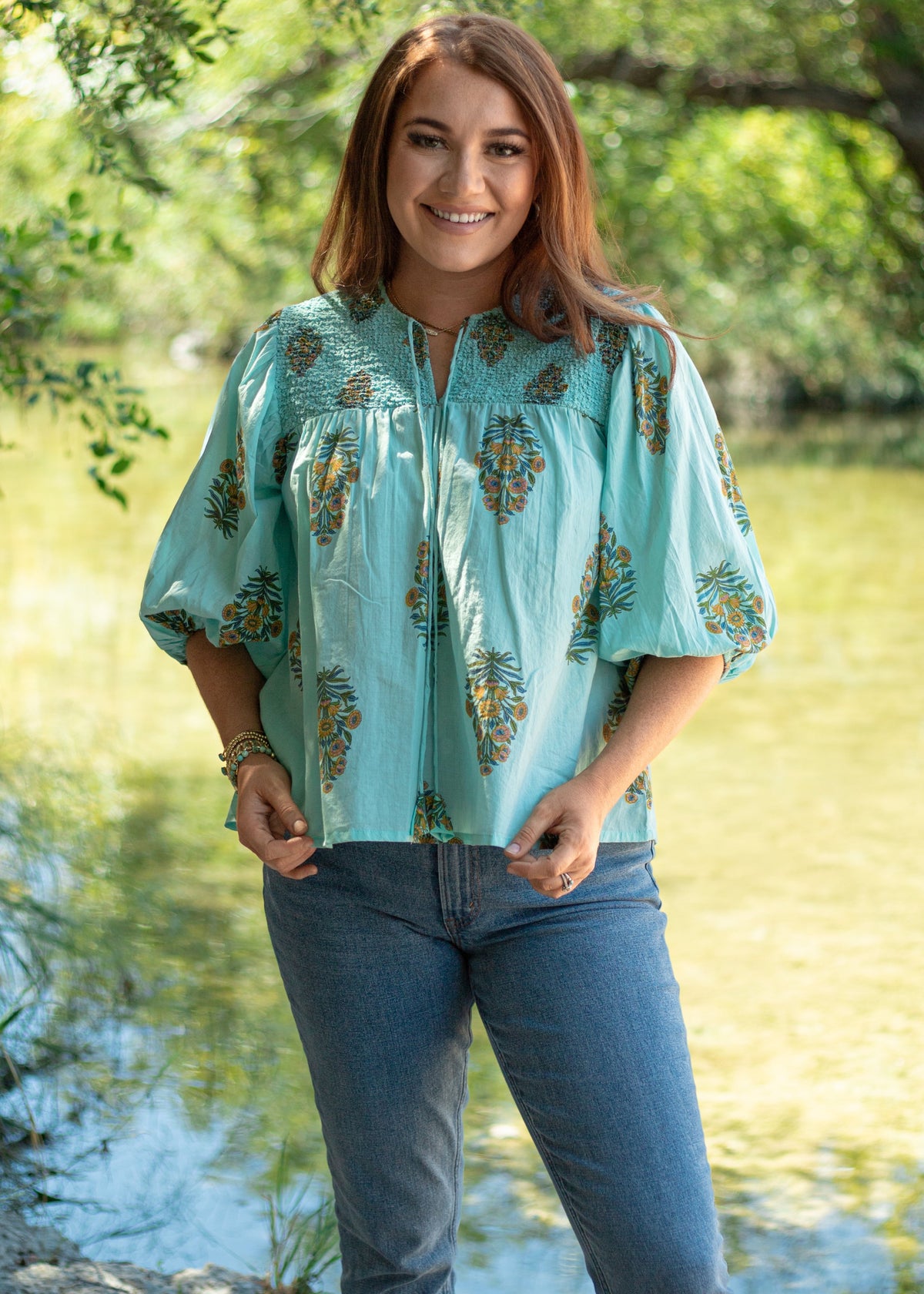 Woman with red hair standing in a front profile position wearing the Ava Top in a lifestyle scene. The focal point of this image is distressed fabric with same graphics along the chest, v-neckline, and tassels. The model paired this top with dark-wash denim jeans. 