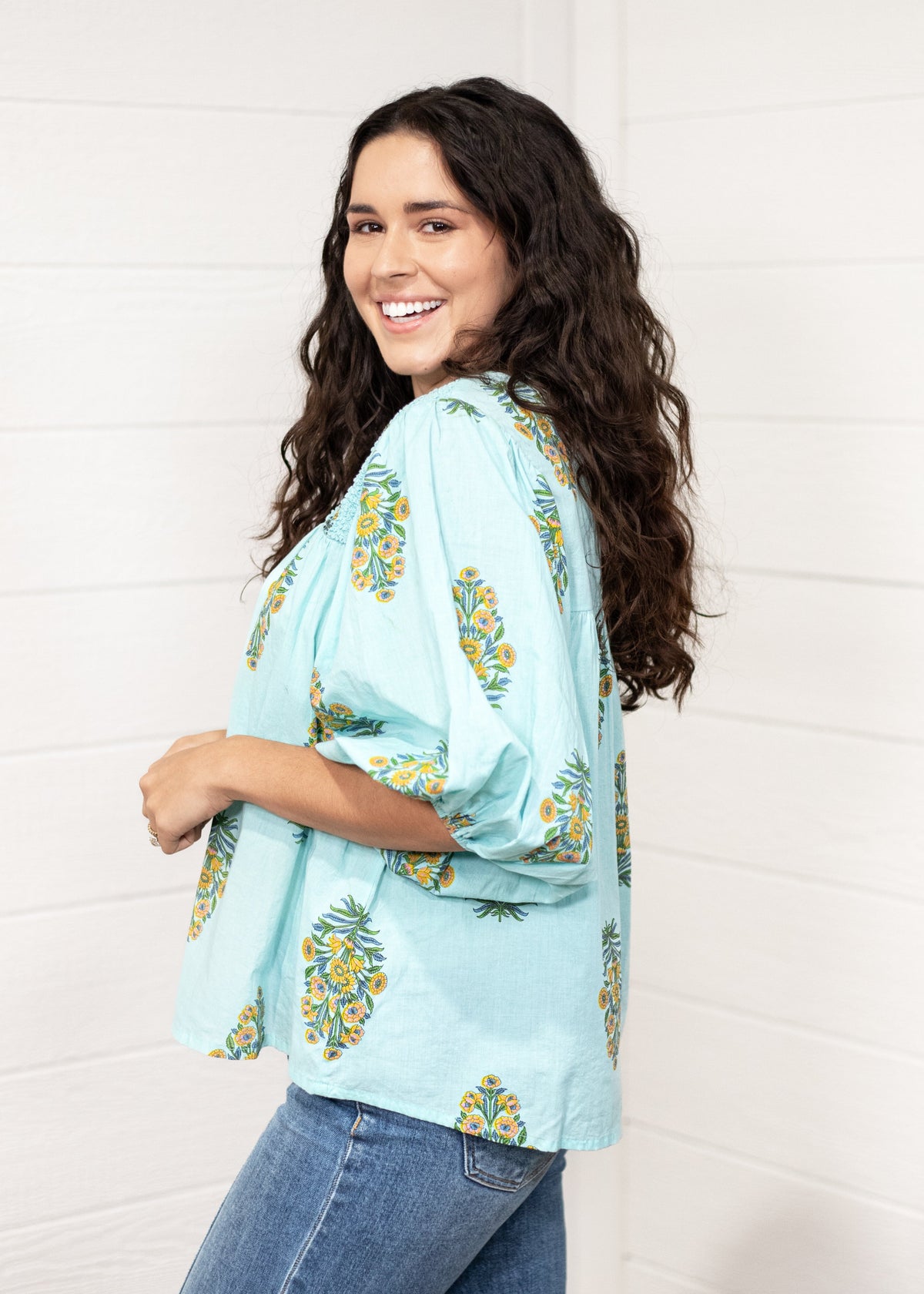 Woman with dark hair standing in a side profile position wearing the Ava Top against a white background. The focal point of this image is to see puffy sleeve length. The model paired this top with dark-wash denim jeans. 