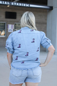 Woman with blonde hair standing in a  back profile position wearing the Bailey Top in a lifestyle scene. The focal point of this image is the purple embroidered boots on back of top as well as back of ruffled sleeves. The model paired this top with light-wash denim shorts. 