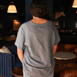 Woman with dark hair standing in a back profile position wearing the Basic Tee in a lifestyle scene. The focal point of this image is the tee's fabric and neckline. The model paired this top with light-wash denim jeans. 
