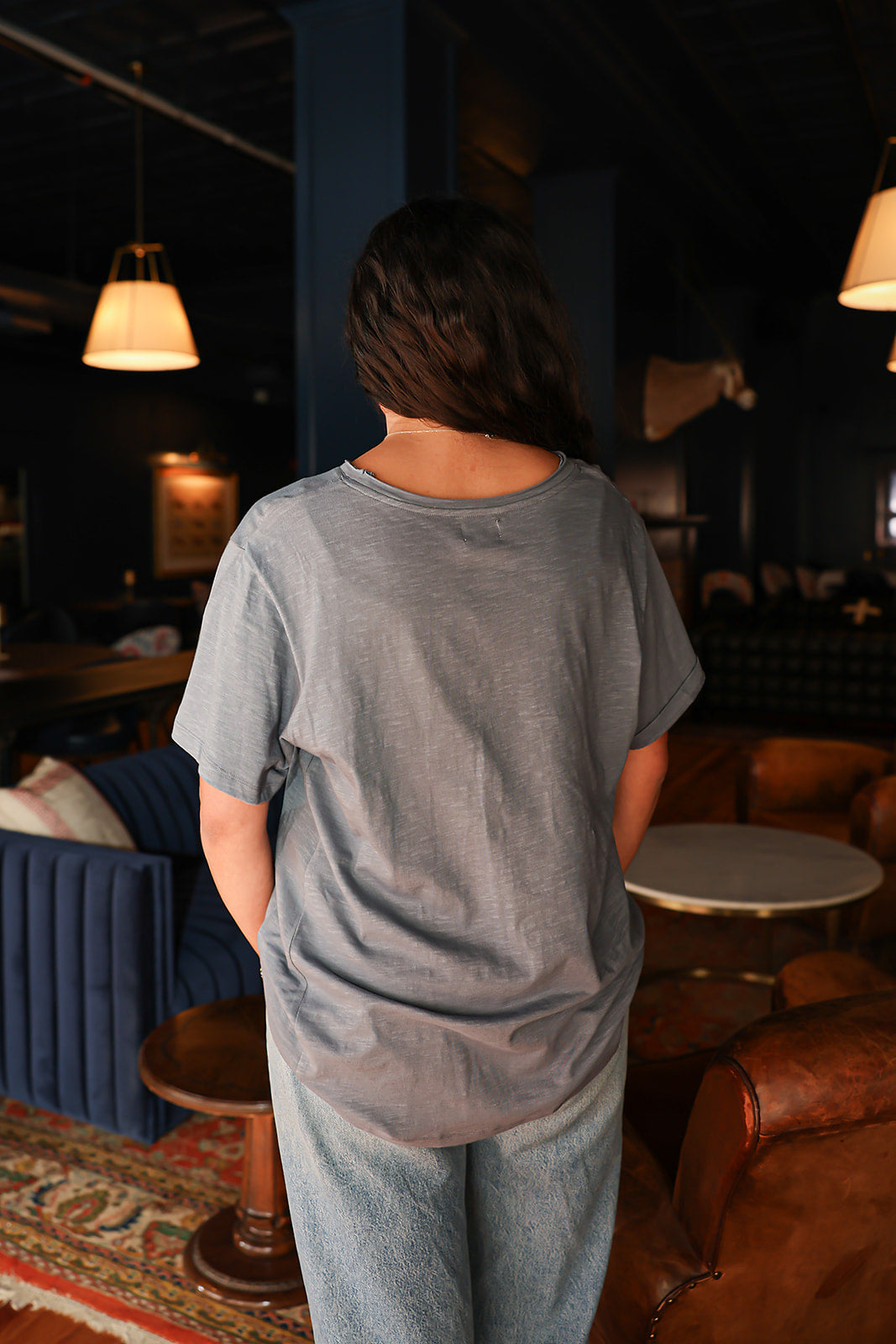 Woman with dark hair standing in a back profile position wearing the Basic Tee in a lifestyle scene. The focal point of this image is the tee's fabric and neckline. The model paired this top with light-wash denim jeans. 