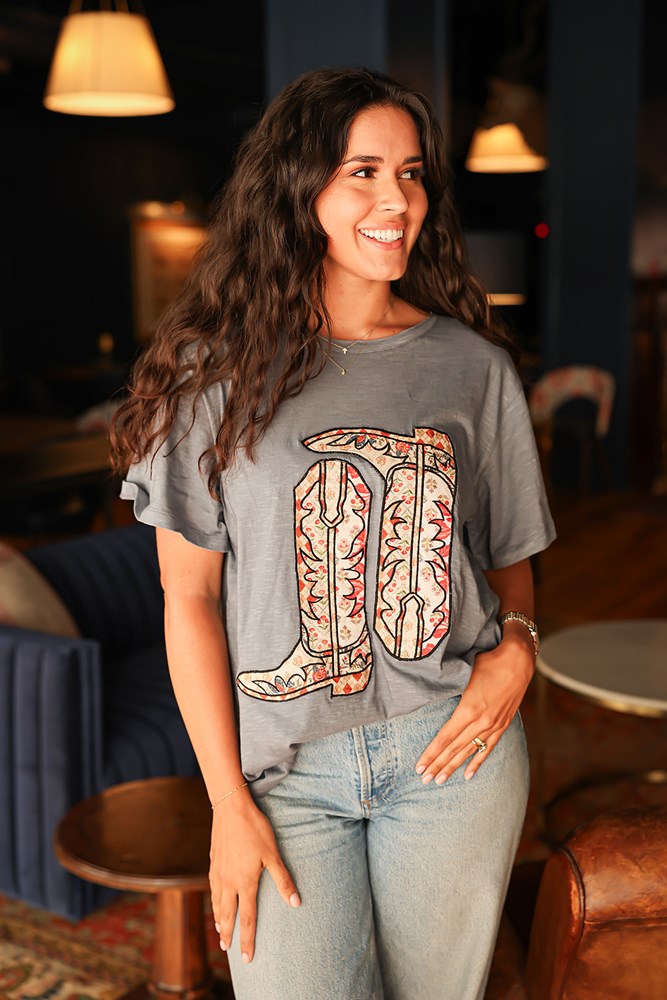 Woman with dark hair standing in a front profile wearing the Basic Tee in a lifestyle scene. The focal point of this image is the embroidered boots on front of top. The model paired this top with light-wash denim jeans. 