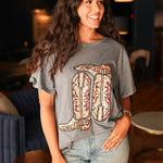 Woman with dark hair standing in a front profile wearing the Basic Tee in a lifestyle scene. The focal point of this image is the embroidered boots on front of top. The model paired this top with light-wash denim jeans. 