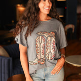 Woman with dark hair standing in a front profile wearing the Basic Tee in a lifestyle scene. The focal point of this image is the embroidered boots on front of top. The model paired this top with light-wash denim jeans. 