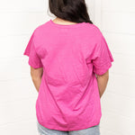 Woman with dark hair standing in a back profile position wearing the Basic Tee against a white background. The focal point of this image is the bubbly pink colored tee. The model paired this tee with light-wash denim shorts.