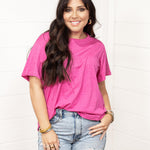 Woman with dark hair standing in a front profile position wearing the Basic Tee against a white background. The focal point for this image is the bubbly pink colored tee. The model paired this tee with light-wash denim shorts.