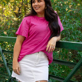 Woman with dark hair standing in a front profile position wearing the Basic Tee in a lifestyle scene. The focal point of this image is the bubbly pink colored tee. The model paired this tee with a white denim skirt. 