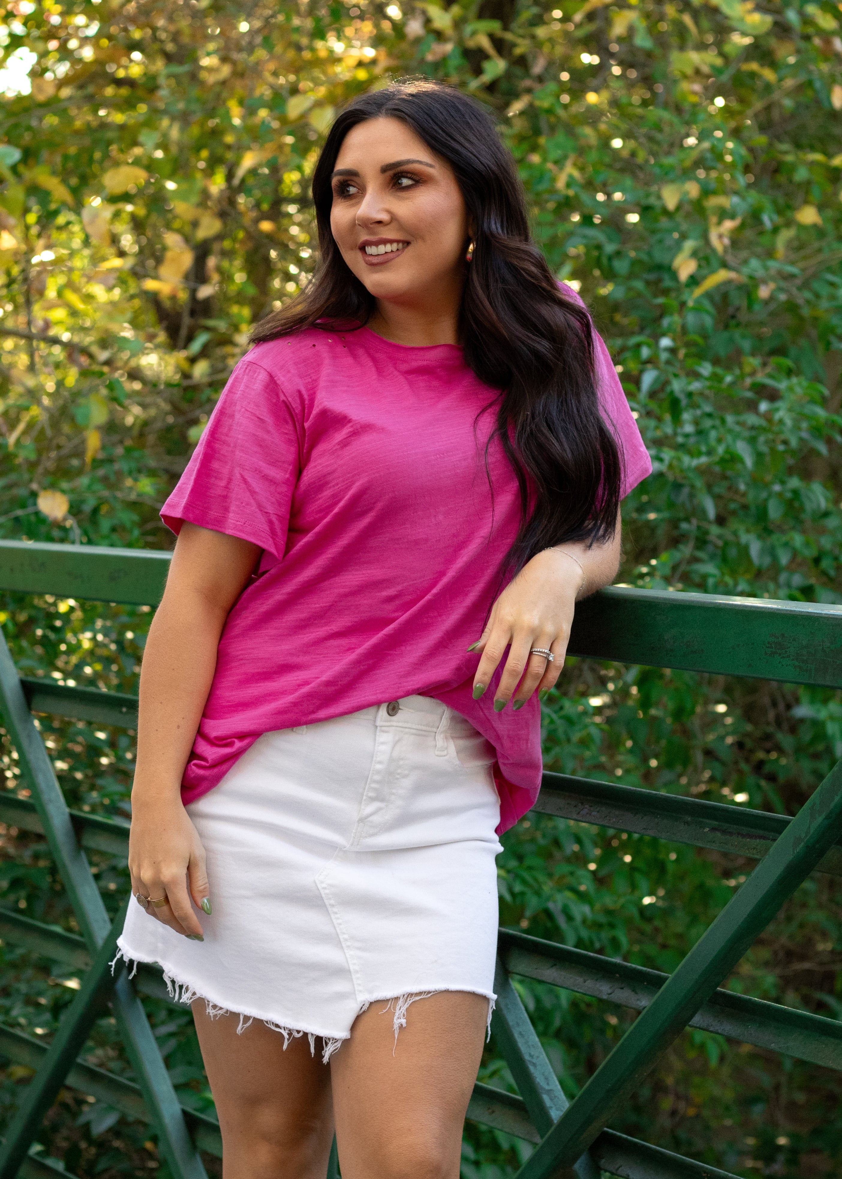 Woman with dark hair standing in a front profile position wearing the Basic Tee in a lifestyle scene. The focal point of this image is the bubbly pink colored tee. The model paired this tee with a white denim skirt. 