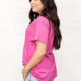 Woman with dark hair standing in a side profile position wearing the Basic Tee against a white background. The focal point for this image is the bubbly pink colored tee. The model paired this tee with light-wash denim shorts. 