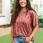 Woman with dark hair standing in a front profile position wearing the Basic Tee in a lifestyle scene. The focal point of this image is the graphics on front of tee. 