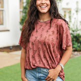 Woman with dark hair standing in a front profile position wearing the Basic Tee in a lifestyle scene. The focal point of this image is the graphics on front of tee. 