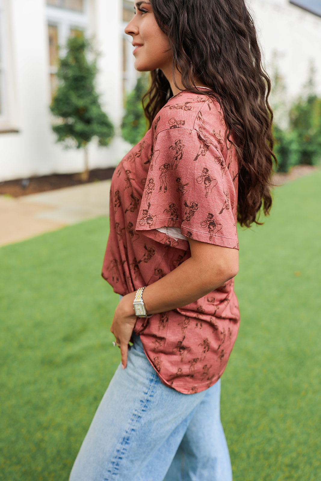 Woman with dark hair standing in a side profile position wearing the Basic Tee in a lifestyle scene. The focal point of this image is the side profile of tee with graphics. The model paired this top with light-wash denim jeans. 