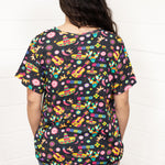 Woman with dark hair standing in a back profile position wearing the Basic Tee against a white background. The focal point of this image is the bright graphics against the black tee (sombreros, guitars, cacti, etc...). The model paired this top with dark-wash denim jeans. 