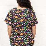 Woman with dark hair standing in a back profile position wearing the Basic Tee against a white background. The focal point of this image is the bright graphics against the black tee (sombreros, guitars, cacti, etc...). The model paired this top with dark-wash denim jeans. 