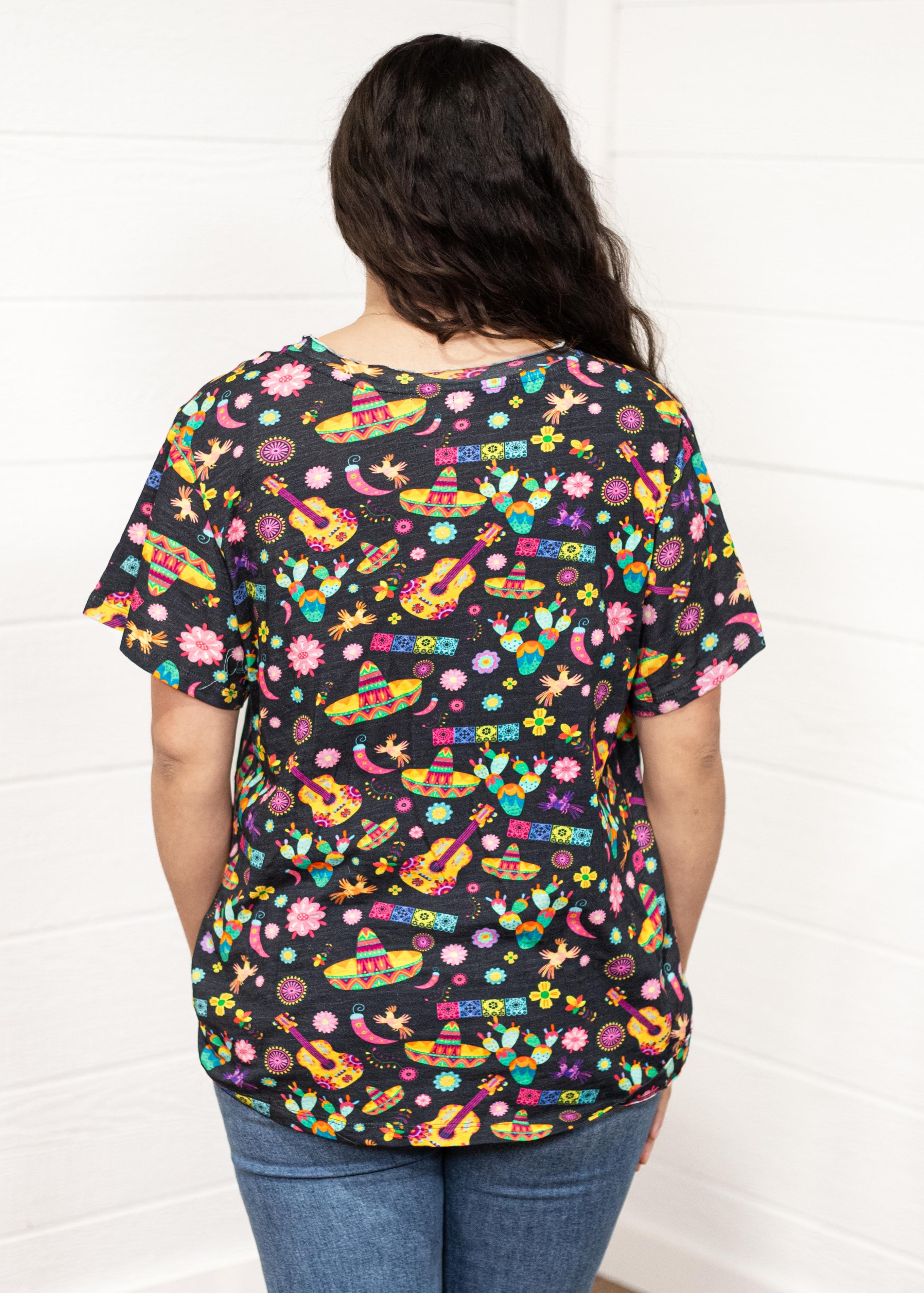 Woman with dark hair standing in a back profile position wearing the Basic Tee against a white background. The focal point of this image is the bright graphics against the black tee (sombreros, guitars, cacti, etc...). The model paired this top with dark-wash denim jeans. 