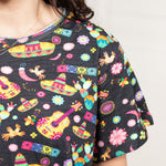 Woman with dark hair standing in a front profile position wearing the Basic Tee against a white background. The focal point of this image is the colorful graphics (sombreros, guitars, cacti, chiles, etc...). 