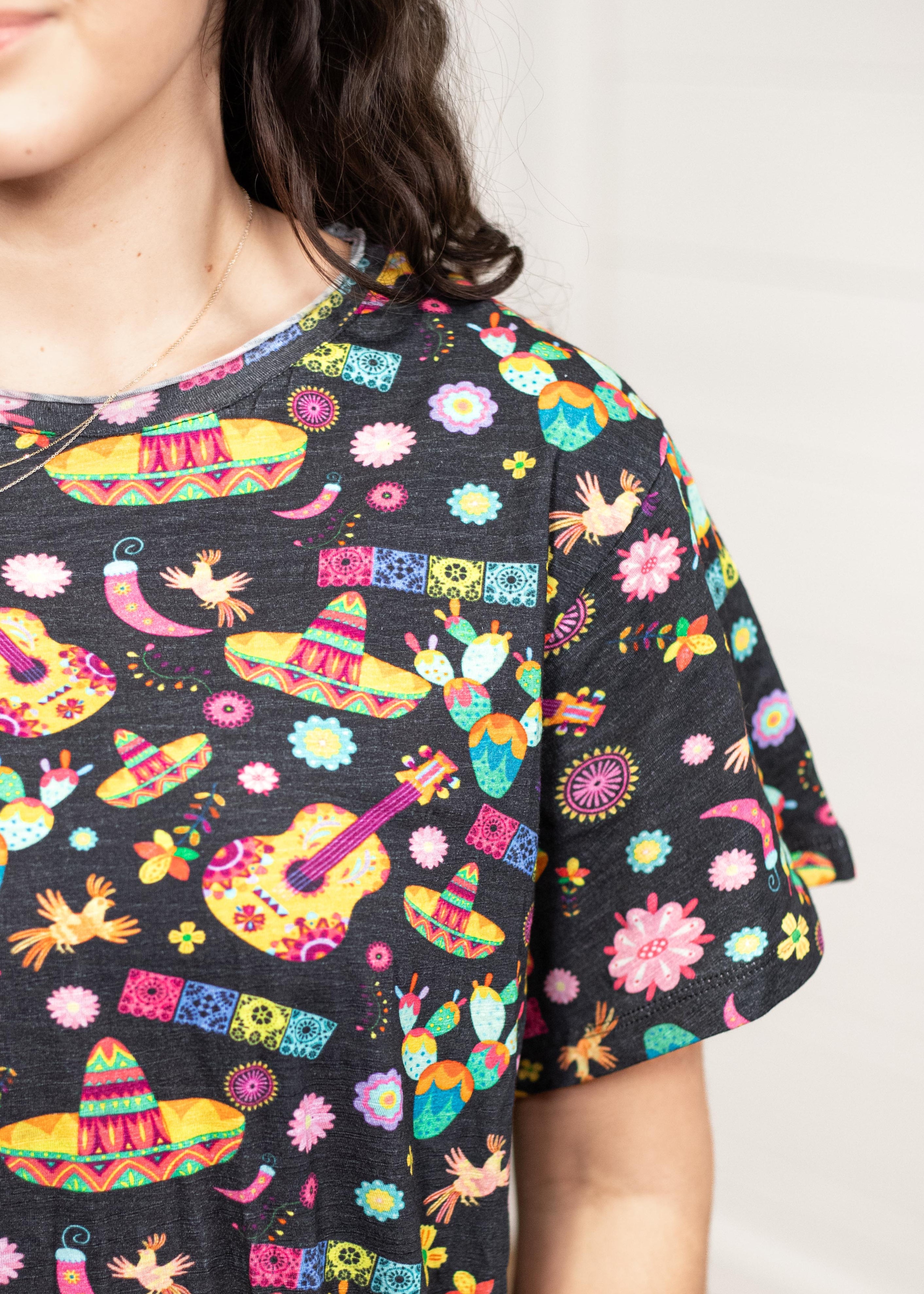 Woman with dark hair standing in a front profile position wearing the Basic Tee against a white background. The focal point of this image is the colorful graphics (sombreros, guitars, cacti, chiles, etc...). 
