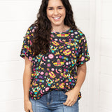 Woman with dark hair standing in a front profile position wearing the Basic Tee against a white background. The focal point of this image is the colorful graphics on tee (sombreros, guitars, cacti, chiles, etc...). The model paired this top with dark-wash denim jeans. 