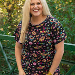 Woman with blonde hair standing in a front profile position wearing the Basic Tee in a lifestyle scene. The focal point of this image is the graphics on tee (sombrero, guitars, cacti, chilies, etc...). The model paired this top with white denim pants. 