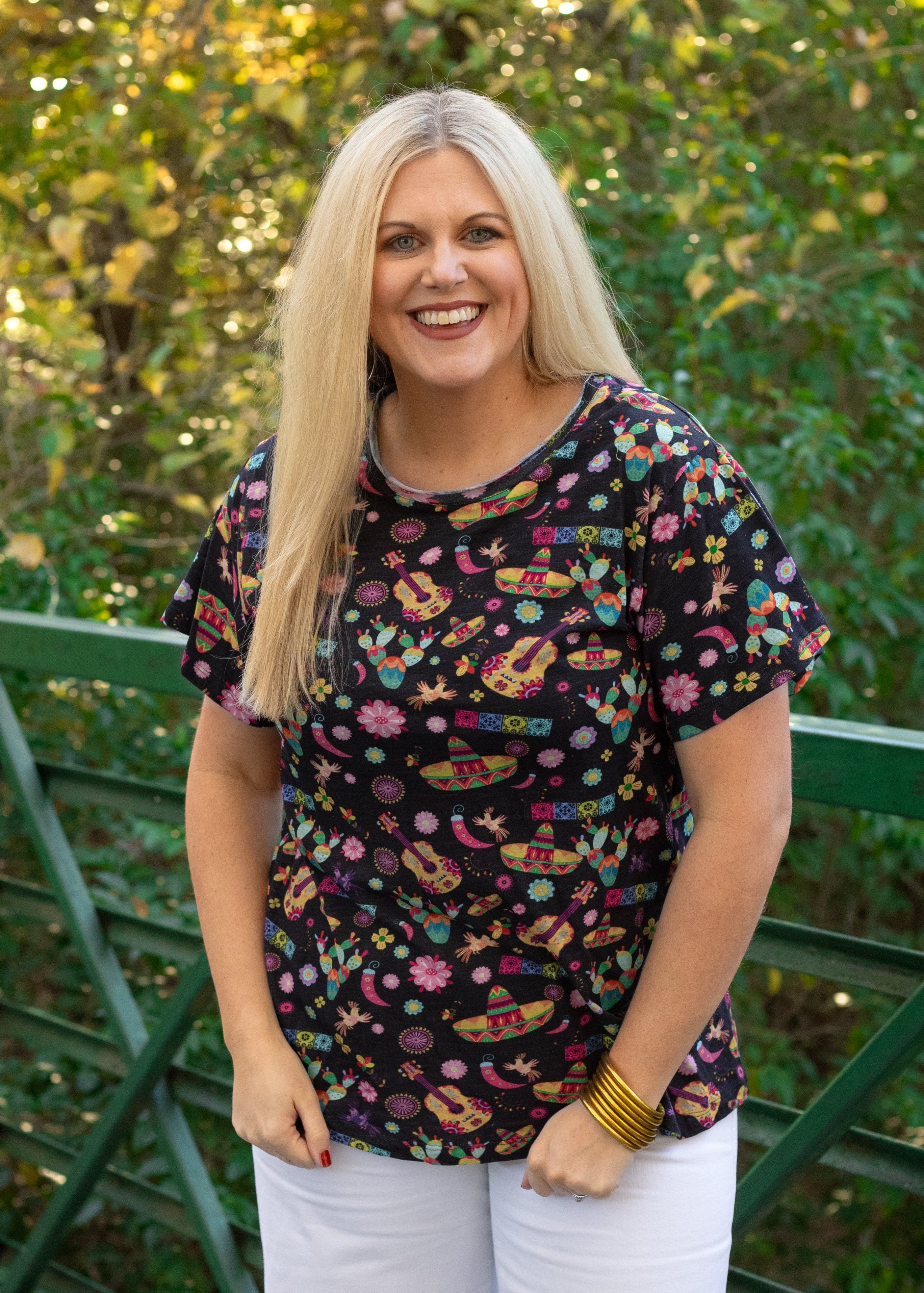 Woman with blonde hair standing in a front profile position wearing the Basic Tee in a lifestyle scene. The focal point of this image is the graphics on tee (sombrero, guitars, cacti, chilies, etc...). The model paired this top with white denim pants. 