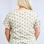 Woman with blonde hair standing in a back profile position wearing the Basic Tee against a white background. The focal point of this image is the pink and green floral pattern. The model paired this tee with light-wash denim shorts. 