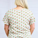 Woman with blonde hair standing in a back profile position wearing the Basic Tee against a white background. The focal point of this image is the pink and green floral pattern. The model paired this tee with light-wash denim shorts. 