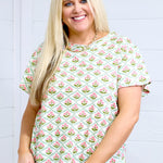 Woman with blonde hair standing in a front profile position wearing the Basic Tee against a white background. The focal point of this image is the pink and green floral graphics. The model paired this top with light-wash denim shorts. 