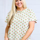 Woman with blonde hair standing in a front profile position wearing the Basic Tee against a white background. The focal point of this image is the pink and green floral graphics. The model paired this top with light-wash denim shorts. 