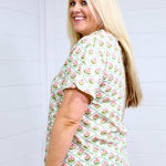 Woman with blonde hair standing in a side profile position wearing the Basic Tee against a white background. The focal point of this image is the pink and green floral graphics. The model paired this top with light-wash denim shorts.