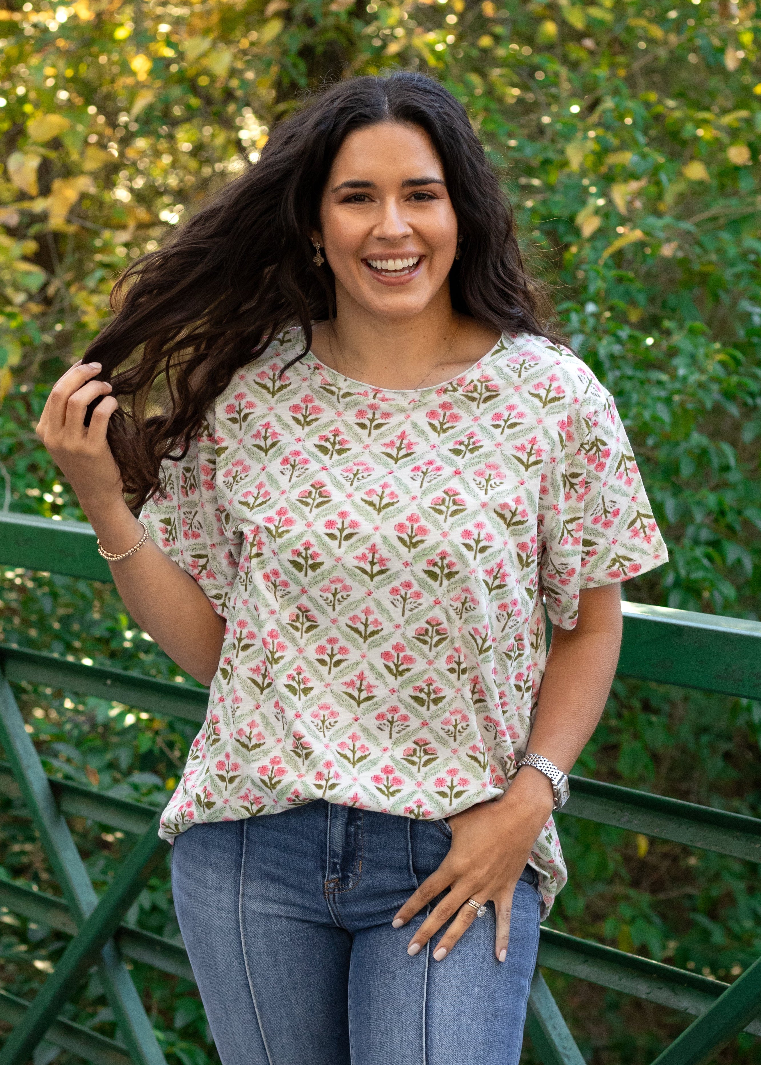 Woman with dark hair standing in a front profile position wearing the Basic Tee in a lifestyle scene. The focal point of this image is the pink and green floral graphics against heavy greenery background. The model paired this top with dark-wash denim jeans. 