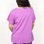 Woman with dark hair standing in a back profile position wearing the Basic Tee against a white background. The focal point of this image is to show back of top and lavender color. The model paired this tee with dark-wash denim jeans.