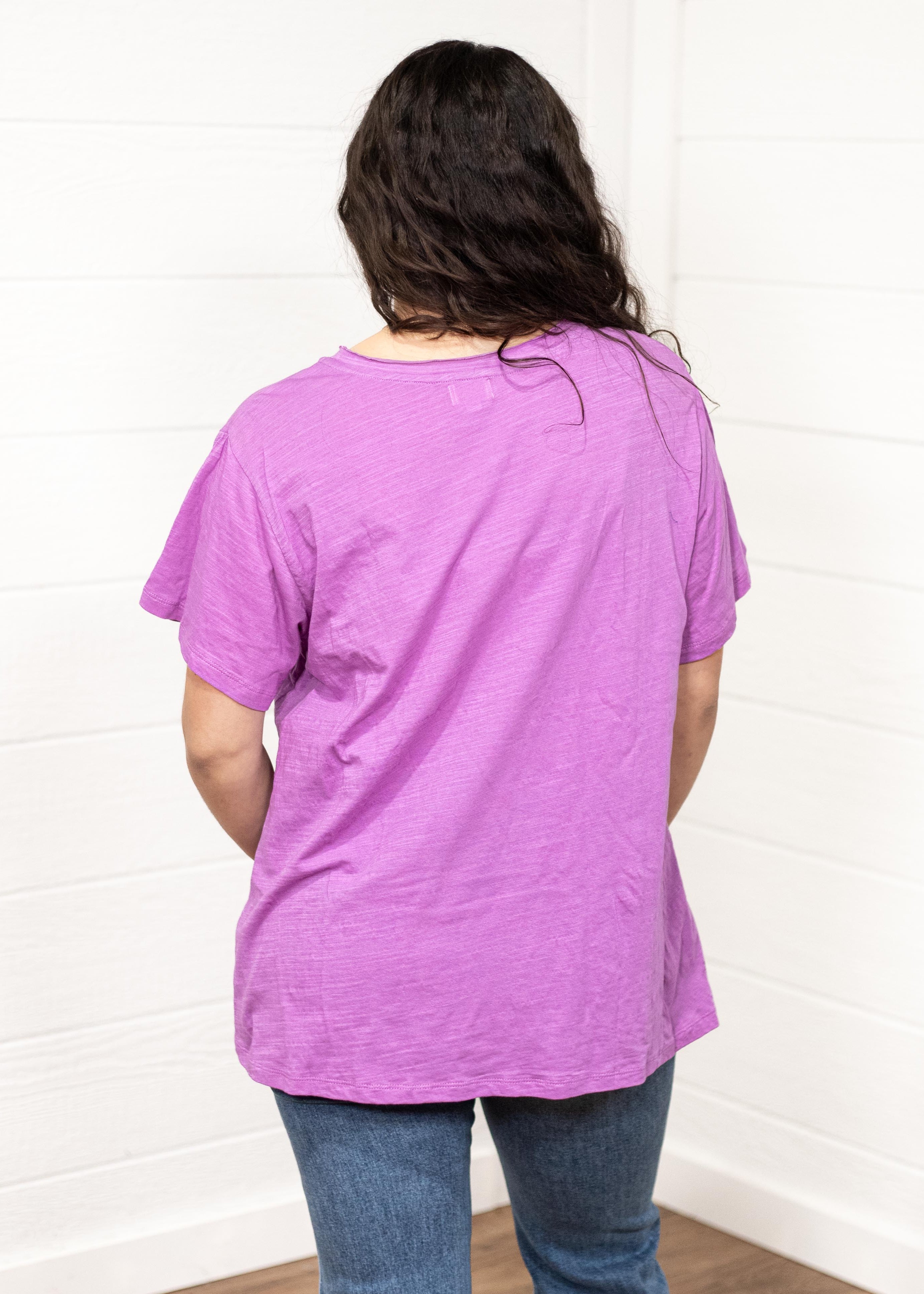 Woman with dark hair standing in a back profile position wearing the Basic Tee against a white background. The focal point of this image is to show back of top and lavender color. The model paired this tee with dark-wash denim jeans.