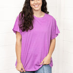 Woman with dark hair standing in a front profile position wearing the Basic Tee against a white background. The focal point of this image is the front of the t-shirt and the lavender color. The model paired this top with dark-wash denim pants. 