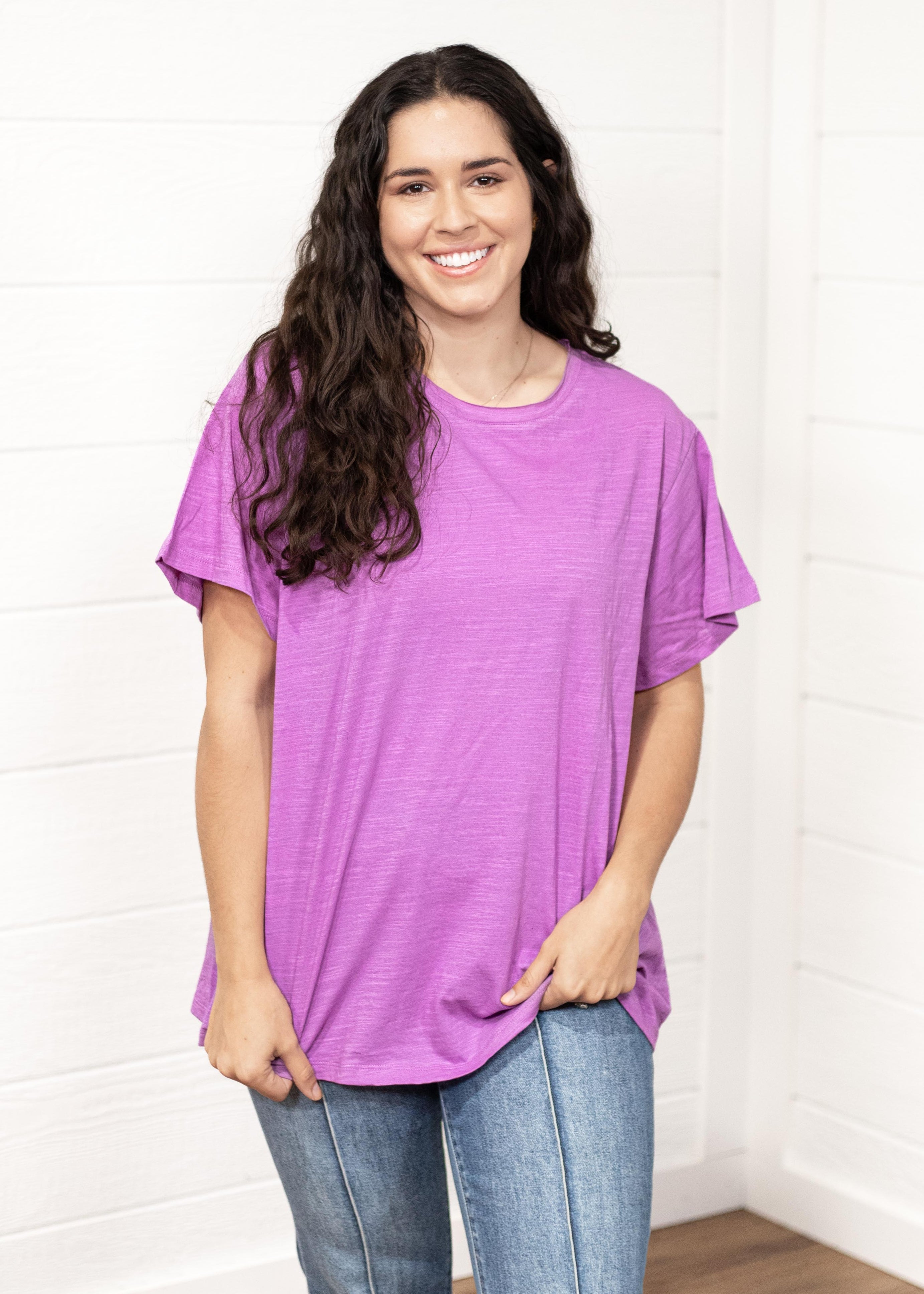 Woman with dark hair standing in a front profile position wearing the Basic Tee against a white background. The focal point of this image is the front of the t-shirt and the lavender color. The model paired this top with dark-wash denim pants. 