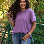 Woman with dark hair standing in a front profile position wearing the Basic Tee in a lifestyle scene. The focal point of this image is the tee in the lavender color against heavy greenery. The model paired this tee with dark-wash denim jeans. 