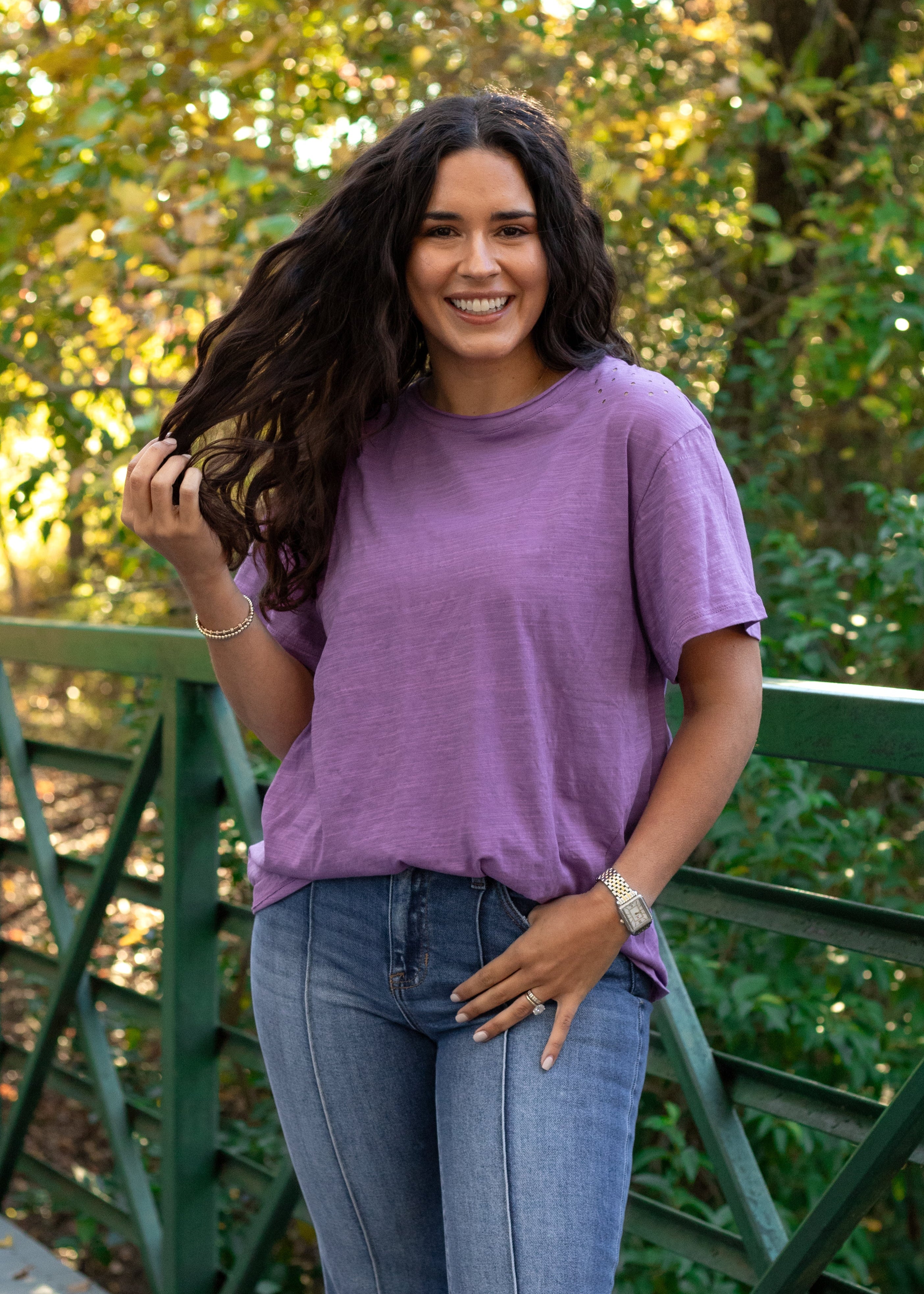 Woman with dark hair standing in a front profile position wearing the Basic Tee in a lifestyle scene. The focal point of this image is the tee in the lavender color against heavy greenery. The model paired this tee with dark-wash denim jeans. 