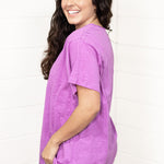 Woman with dark hair standing in a side profile position wearing the Basic Tee against a white background. The focal point of this image is the side of tee and lavender color. The model paired this tee with dark-wash denim pants.