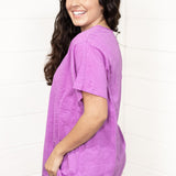 Woman with dark hair standing in a side profile position wearing the Basic Tee against a white background. The focal point of this image is the side of tee and lavender color. The model paired this tee with dark-wash denim pants.