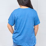 Woman with dark hair standing in a back profile position wearing the Basic Tee against a white background. The focal point of this image is the blue t-shirt. The model paired this tee with dark-wash denim shorts. 