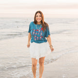 Woman with dark hair standing in a front profile position wearing the Basic Tee in a lifestyle scene. The focal point of this image is the front embroidery of the tee. The model paired this top with a white skirt.