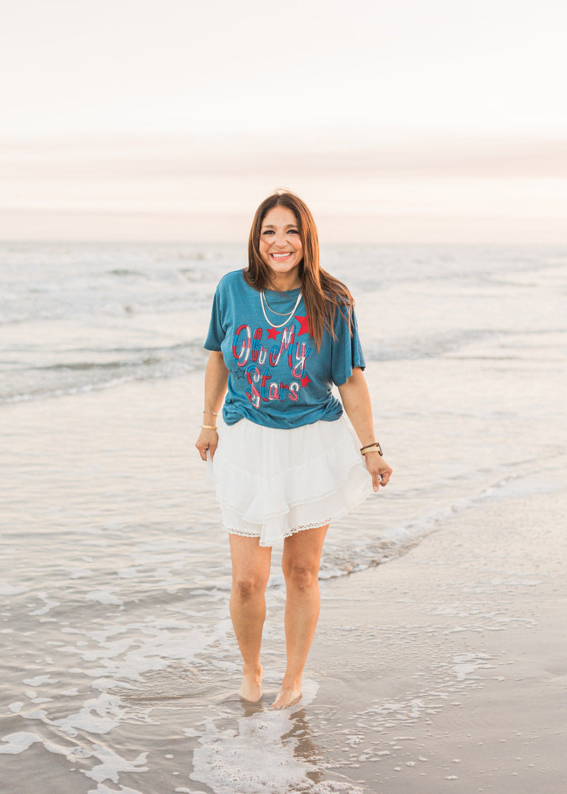 Woman with dark hair standing in a front profile position wearing the Basic Tee in a lifestyle scene. The focal point of this image is the front embroidery of the tee. The model paired this top with a white skirt.