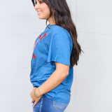 Woman with dark hair standing in a side profile position wearing the Basic Tee against a white background. The focal point of this image is the red embroidered stars peeking from front of top and length at waist and sleeve. The model paired this top with dark-wash denim shorts. 