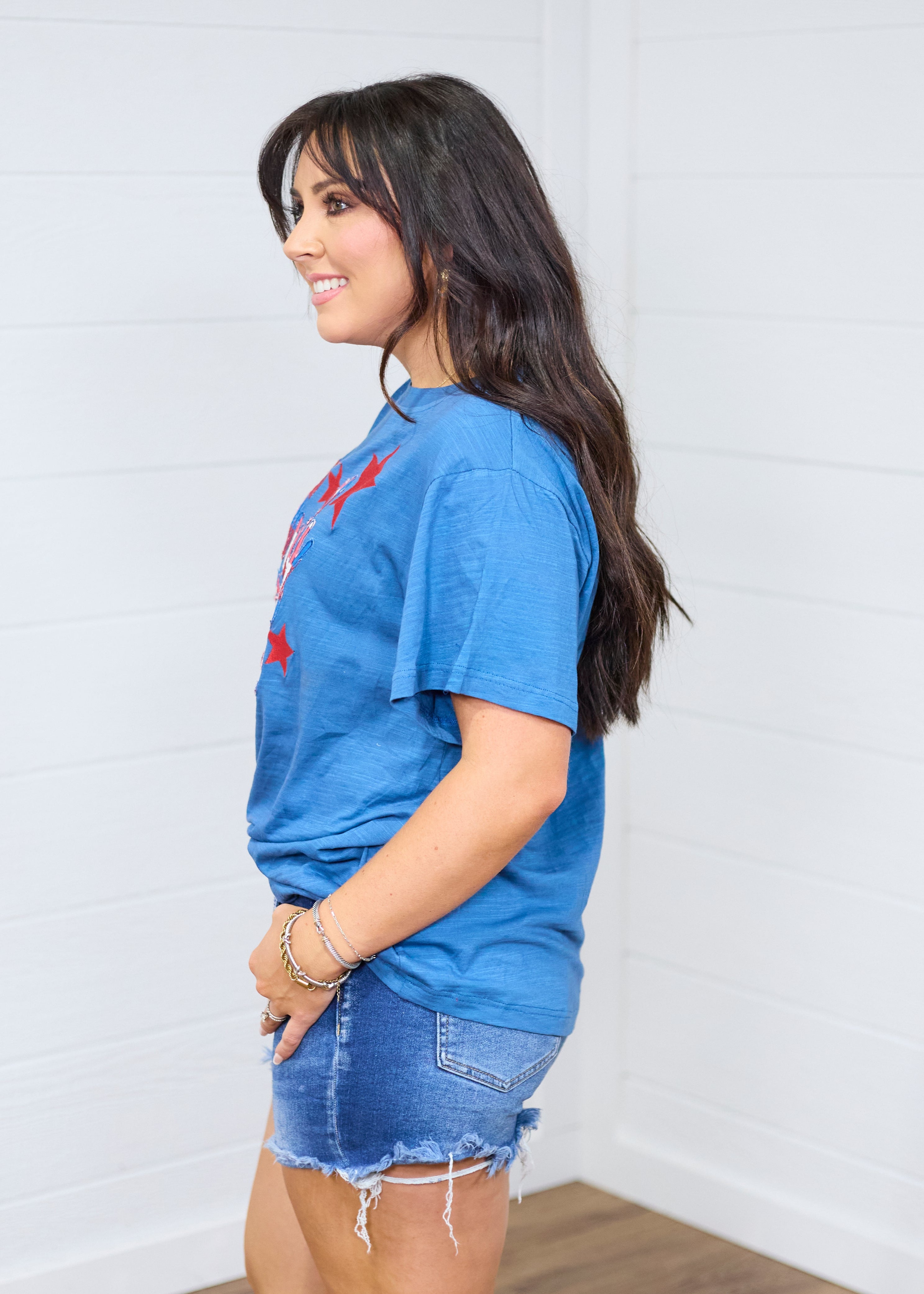 Woman with dark hair standing in a side profile position wearing the Basic Tee against a white background. The focal point of this image is the red embroidered stars peeking from front of top and length at waist and sleeve. The model paired this top with dark-wash denim shorts. 
